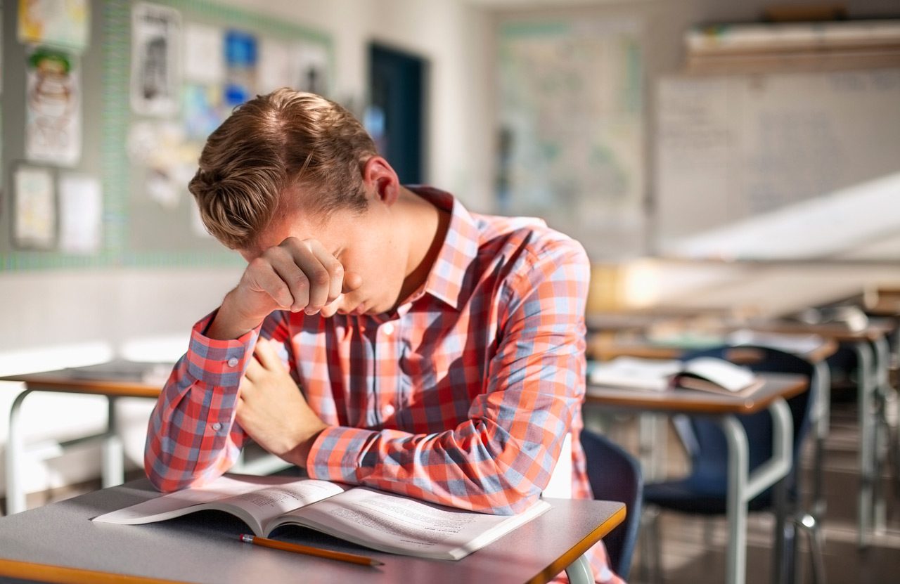 Stressed-male-student-sitting-with-book-at-desk